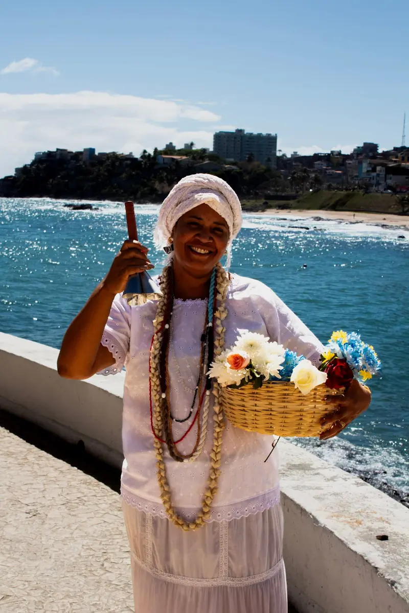 Mãe Iara sorrindo com adjá e cesta de flores à beira-mar